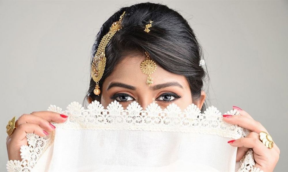 A woman partially covers her face with a delicate white lace fabric while revealing expressive eyes highlighted by dark eyeliner. Her beautifully styled hair is adorned with traditional gold jewelry including a maang tikka on her forehead. She holds the fabric with red manicured nails that add color to her soft look against a neutral background, showcasing elegance and cultural elements present in wedding traditions.