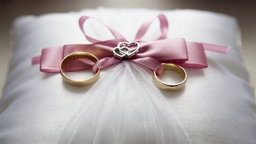Two gold wedding rings resting on a white satin pillow adorned with delicate tulle and topped with a pink ribbon bow. The bow features interlocking silver hearts at the center, symbolizing love and commitment. Soft textures create an elegant look, showcasing the romance of weddings.