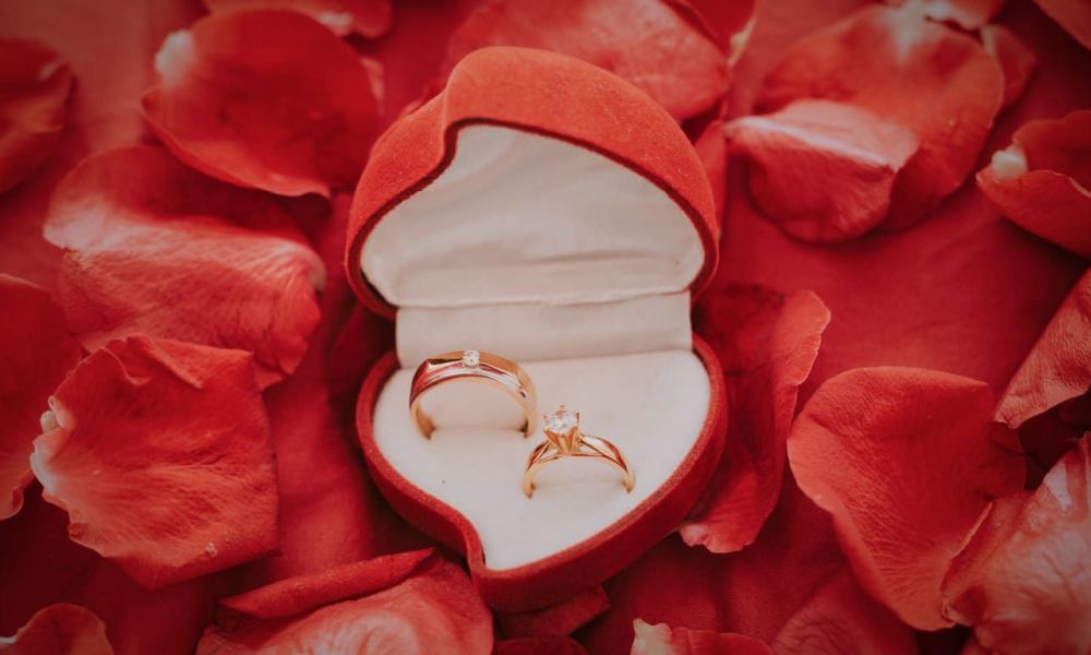 A beautiful open red velvet heart-shaped jewelry box displays two stunning gold rings, one adorned with a diamond. The box is beautifully arranged on a surface with scattered red rose petals adding elegance to the scene. Soft lighting creates a romantic atmosphere, perfect for engagements or weddings.