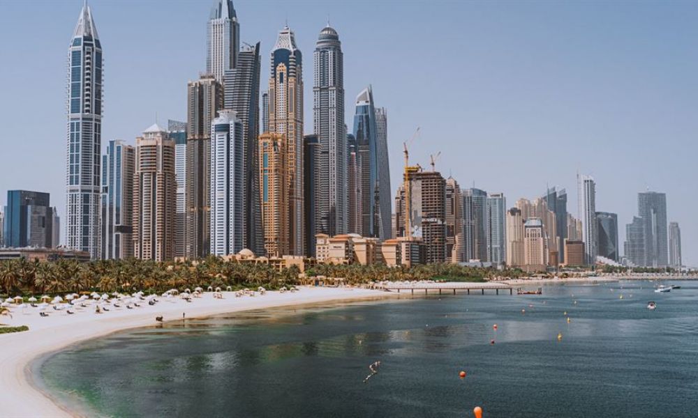 A breathtaking view of Dubai's skyline amidst clear blue skies showcases modern skyscrapers and serene palm-lined beaches. The sun-kissed waters reflect elegant hotels that define luxury living, creating an inviting atmosphere for couples looking to celebrate their love here. Buoys add colorful pops around the tranquil coastline surrounded by life’s energy and beauty. This image embodies romance and excitement for those dreaming of a perfect wedding day in Dubai.