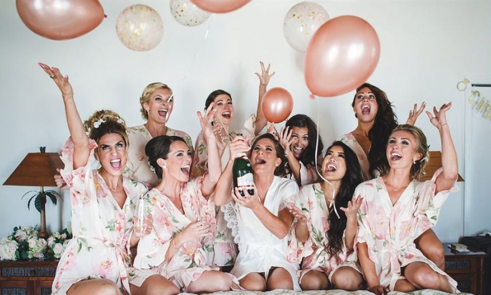A joyful group of women dressed in matching floral robes celebrate together indoors with raised hands and big smiles. One woman holds a champagne bottle while pink and cream balloons float above them adding festivity. The backdrop features a bride-to-be banner enhancing the celebratory atmosphere typical of bridal showers or pre-wedding events. The overall scene radiates excitement, happiness, and camaraderie among friends preparing for a wedding celebration.