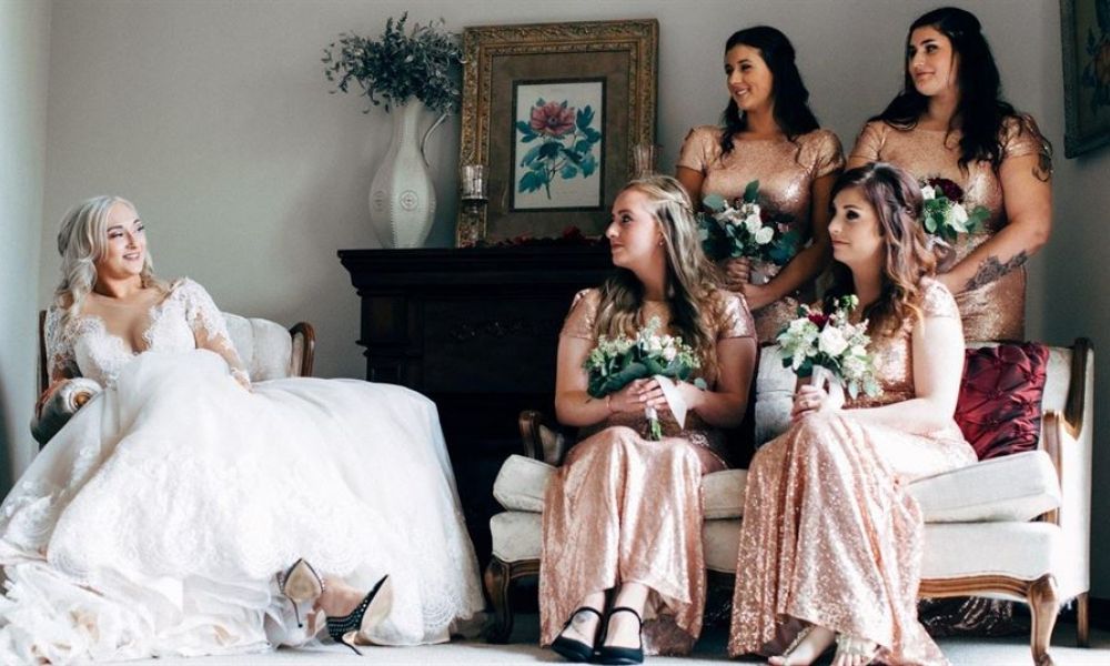 Image of a happy bride in a white gown sitting gracefully on a chair surrounded by her bridesmaids in shimmering rose gold dresses, all smiling together before their wedding ceremony in an elegantly decorated room with soft natural light illuminating their joy.