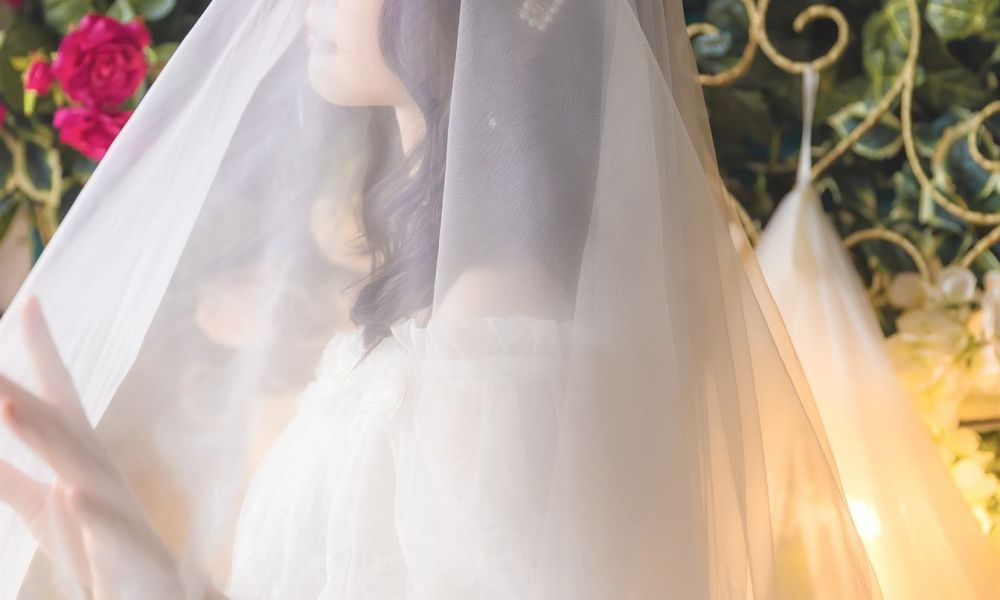 A bride stands in a lush garden filled with colorful flowers and buzzing greenery. She wears a stunning off-the-shoulder white lace dress that billows gently around her. Delicate wax-like pearls adorn her styled hair, which cascades softly over her shoulders. The enchanting soft light creates an ethereal ambiance accentuated by lovely wrought iron details in the background, adding elegance to this romantic scene perfect for weddings.