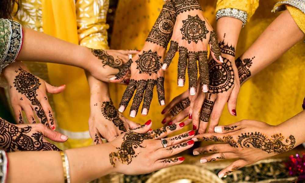 A mesmerizing display of hands adorned with intricate henna patterns, showcasing rich floral and geometric designs. The hands are arranged together, symbolizing unity and celebration. Brightly colored outfits in shades of yellow and green hint at a festive occasion, likely connected to a wedding or cultural festivity. This image encapsulates the artistry and significance of henna in joyful celebrations.
