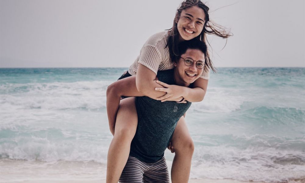 A joyful couple experiences playful moments on the beach. The man gives the woman a piggyback ride as waves gently crash behind them. Both wear casual summer outfits while smiling under a partly cloudy sky. The scene captures their love and fun by the ocean.