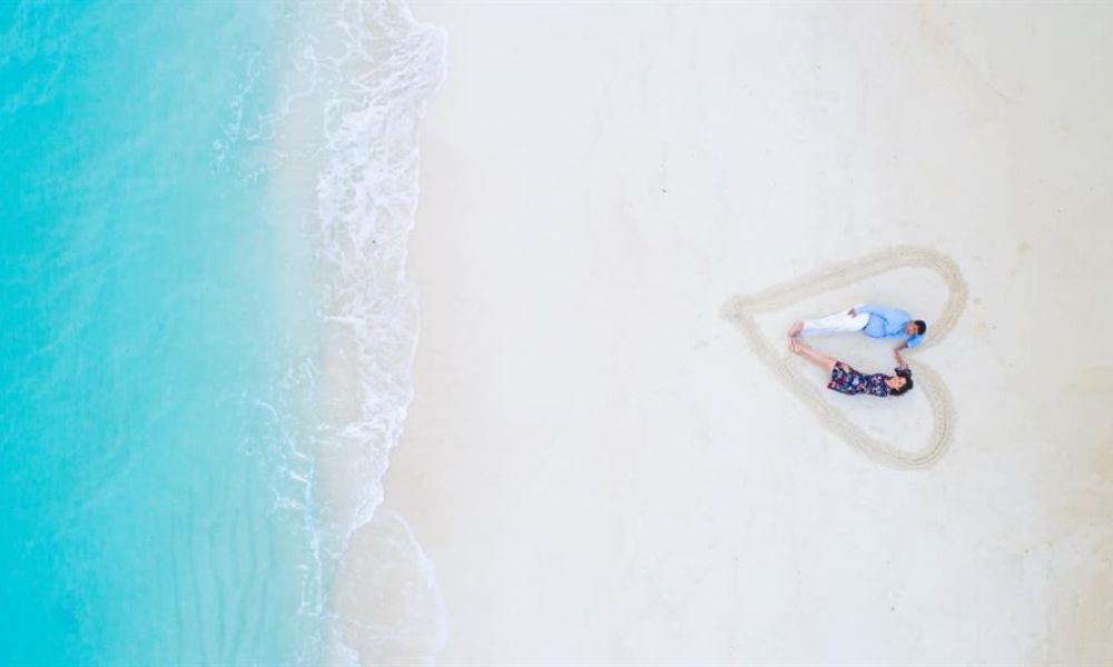 Marrying In Seychelles As A Uae Expat Heres 3 Things To Keep In Mind De1Ffd67 | Marrying In Seychelles As A Uae Expat; Here'S 3 Things To Keep In Mind | Due To The Tideous Documentation, Paperwork And Long Wait For Dates At The Dubai Court; Many Expats Are Now Choosing To Get Married In Seychelles. A couple lying together forms a heart shape on a sandy beach with turquoise ocean waves nearby. The man wears a light blue shirt and white pants while the woman is dressed in a dark floral dress. They appear relaxed and happy surrounded by powdery sand under bright sunlight.