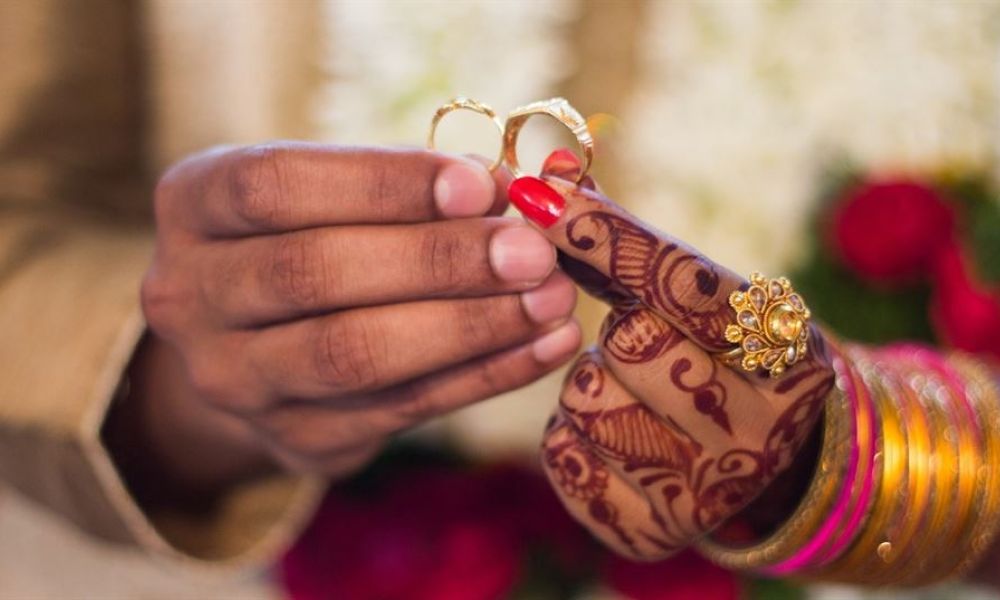 The image displays two hands holding gold rings against a blurred floral background. One hand shows colorful bangles and intricate henna designs while presenting a ring delicately between the thumb and forefinger. This imagery symbolizes love and commitment within a wedding context.