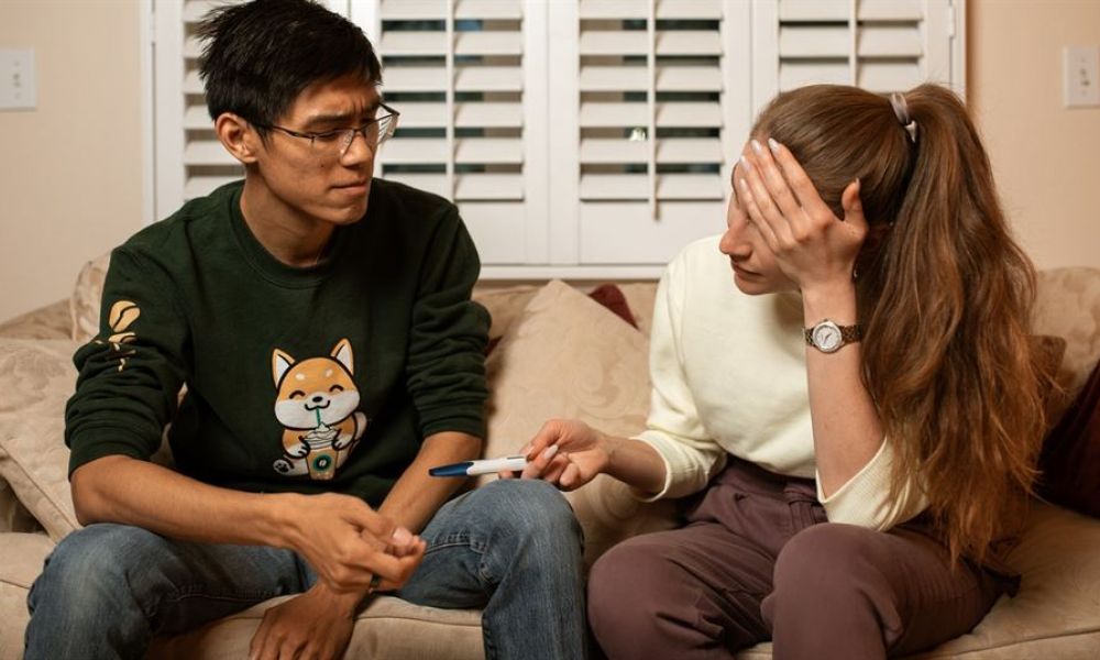 A couple sitting on a beige couch looks concerned as they discuss something important. The man wears a green sweatshirt and glasses while gesturing with his hand. The woman is in a white sweater, holding her forehead with one hand and showing him a pregnancy test with the other hand. They appear intimate and emotional, surrounded by soft lighting from window shutters behind them.