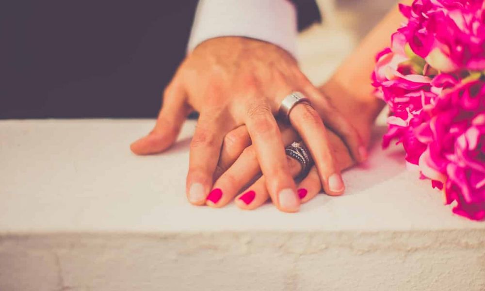 A romantic scene featuring two clasped hands adorned with rings resting on a white surface. One hand has bright red polished nails while next to them lies a bouquet of vibrant pink flowers. The image captures themes of love and commitment in an intimate setting with warm lighting that emphasizes the joy of getting married. This visually speaks to couples considering their wedding options in Dubai and Abu Dhabi for their big day.