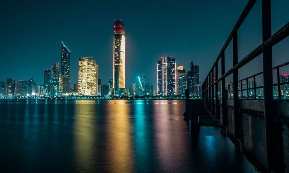 The image features a breathtaking nighttime view of Abu Dhabi with illuminated skyscrapers reflecting on water. It shows a vibrant cityscape filled with lights creating an enchanting atmosphere. A walkway extends over the tranquil surface, leading into this lively scene where modernity meets natural beauty under deep blue skies accented by warm yellow lights.