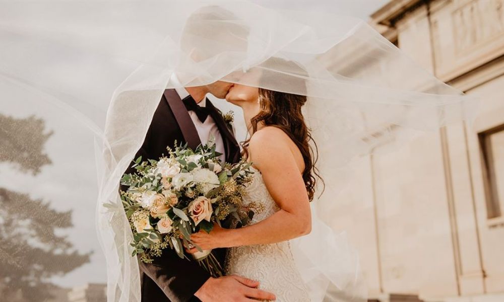 A romantic couple sharing a kiss on their wedding day outdoors. The bride wears a beautiful lace gown while holding pastel flowers. Her flowing veil glows softly in the sunlight, creating a dreamy effect around them. The groom looks dapper in his classic suit amidst an elegant architectural backdrop that indicates their wedding venue. This image captures the intimate joy of their celebration, making it perfect for couples dreaming of their own special moment in Dubai.