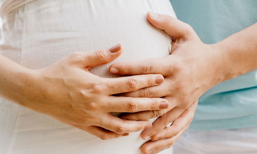 The image features a pregnant person in a white dress being embraced by another individual in a teal shirt and white shorts. Both hands are gently placed on the round belly, symbolizing care and connection. The background softly blurs, highlighting this intimate moment shared between them, bathed in warm natural lighting.