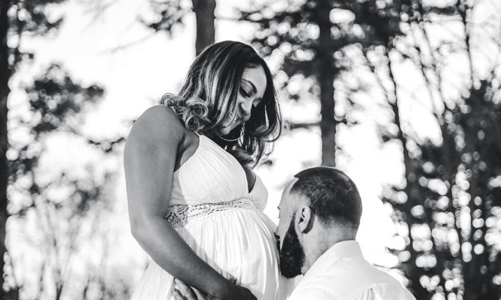 The image shows a loving couple outdoors surrounded by trees. A pregnant woman in a flowing white dress gazes down at her partner who is kneeling or crouching before her. He tenderly kisses her baby bump conveying warmth and affection between them as they anticipate their future together.