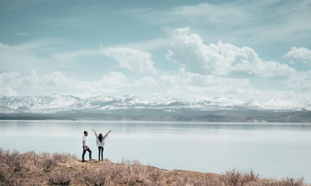 A beautiful image shows a serene view of two people by calm water. They are surrounded by fluffy clouds and mountains. One person raises their arms joyfully while the other admires the scenery. This captures a peaceful moment shared in nature, reflecting happiness and love in an enchanting setting.