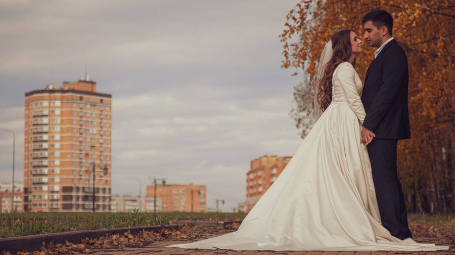 A bride and groom share a loving moment outdoors on a paved path surrounded by vibrant autumn trees. The bride wears a long white gown with a veil while the groom is elegantly dressed in a dark suit. Tall apartment buildings form an urban backdrop against the serene scene filled with golden leaves covering the ground, capturing emotion and warmth on this dreamy day.