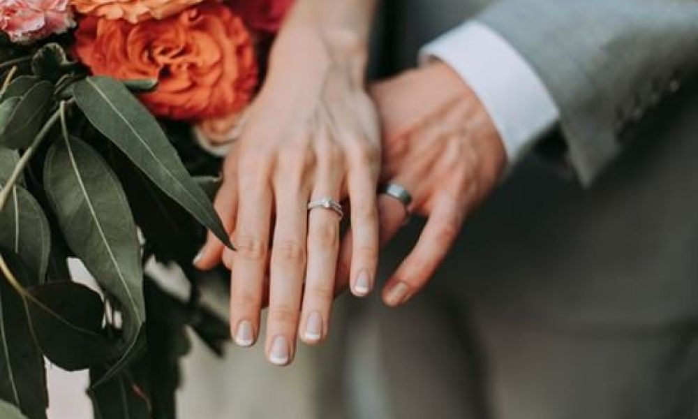 The image shows two hands wearing beautiful wedding rings together with colorful flowers in the background. One hand has part of a grey suit sleeve visible, highlighting an intimate moment of commitment between the couple. This romantic scene invokes love and celebration, perfect for weddings or engagements.