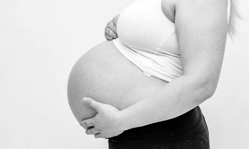 A black-and-white photograph shows a pregnant woman’s side profile wearing a fitted white tank top and dark skirt. Her right arm cradles her baby bump while her left arm supports the bottom of her belly. The light-colored background emphasizes her form, capturing a serene moment that highlights themes of motherhood and anticipation without showing her face or surroundings.