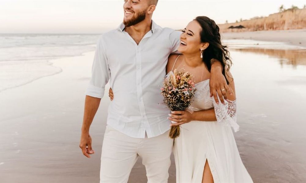 not a big fan of cold weather plan your uae destination wedding in the tropical paradise of seychelles 096c9819 | Not a Big Fan of the Cold Weather? Plan your UAE Destination Wedding in the Tropical Paradise of Seychelles! | The tropical paradise of Seychelles bangs in the Indian ocean which is a group of over 100 small islands. This place has also become synonymous with island life, sparkling beaches, romantic honeymoons, relaxing holidays, wedding paradise, and more. The attraction of having a wedding in Seychelles lies within its stunning, unspoilt, and pristine beauty. The beaches that Seychelles has to offer are second to none in the whole World. That’s why planning your dream destination wedding in this paradise is a perfect way to make your big day even more special. A couple stands happily on a sandy beach as waves gently lap at the shore. The man wears a white shirt with light pants while his partner is dressed in a flowing white gown adorned with lace sleeves. She holds a beautiful bouquet of flowers. They smile at each other, basking in warm sunlight against an idyllic ocean backdrop filled with calm waves and distant cliffs, creating an atmosphere of joy and tranquility perfect for their wedding day celebration.