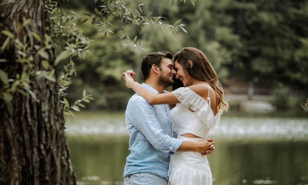 A loving couple embraces by a serene lake, creating a romantic scene surrounded by lush greenery. The man wears a light blue shirt while the woman looks beautiful in her white lace dress, symbolizing love and commitment. This idyllic outdoor setting enhances their affectionate moment together, portraying warmth and tranquility that resonates with every couple's special day.