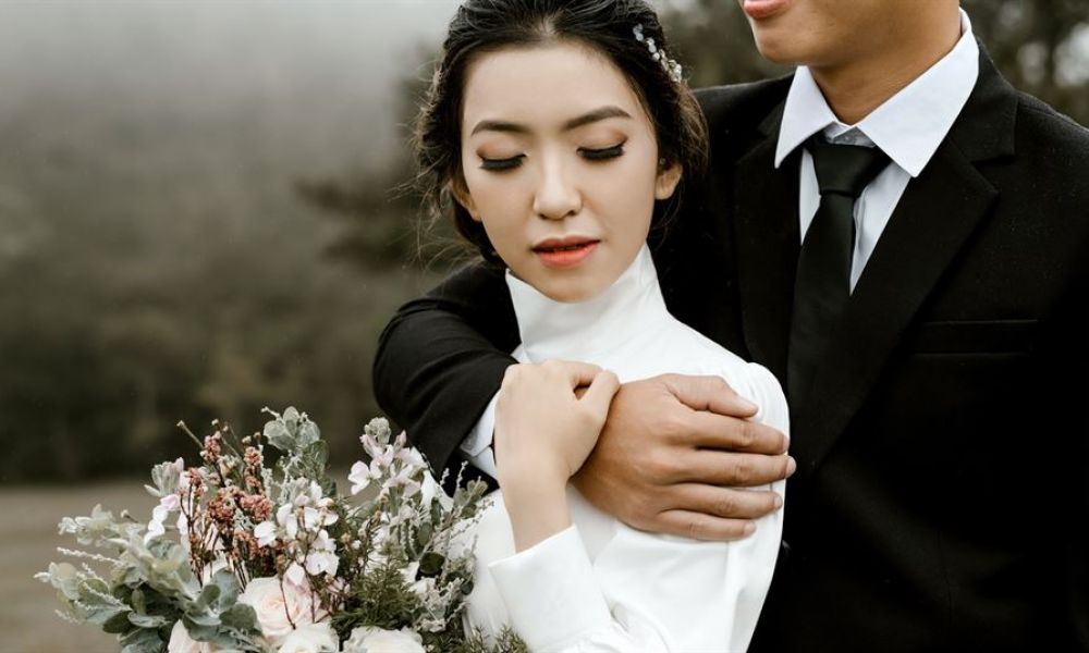 A happy couple dressed in a white wedding dress and a black suit embrace each other tenderly outdoors, surrounded by soft greenery. The woman smiles peacefully while holding a bouquet of pink and white flowers. Their romantic moment is captured beautifully with a dreamy blurred background, creating an atmosphere filled with love and joy perfect for weddings in Dubai.