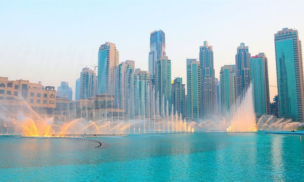 A beautiful sunset over the Dubai skyline showcases tall modern buildings reflecting on water pools. A colorful fountain show lights up the scene, creating a magical atmosphere perfect for weddings. The sparkling waters dance harmoniously as skyscrapers stand proudly in the background, displaying Dubai's architectural wonders. Ideal for couples dreaming of getting married or celebrating their love amidst stunning views, this vision captures romance and excitement that awaits you in Dubai.