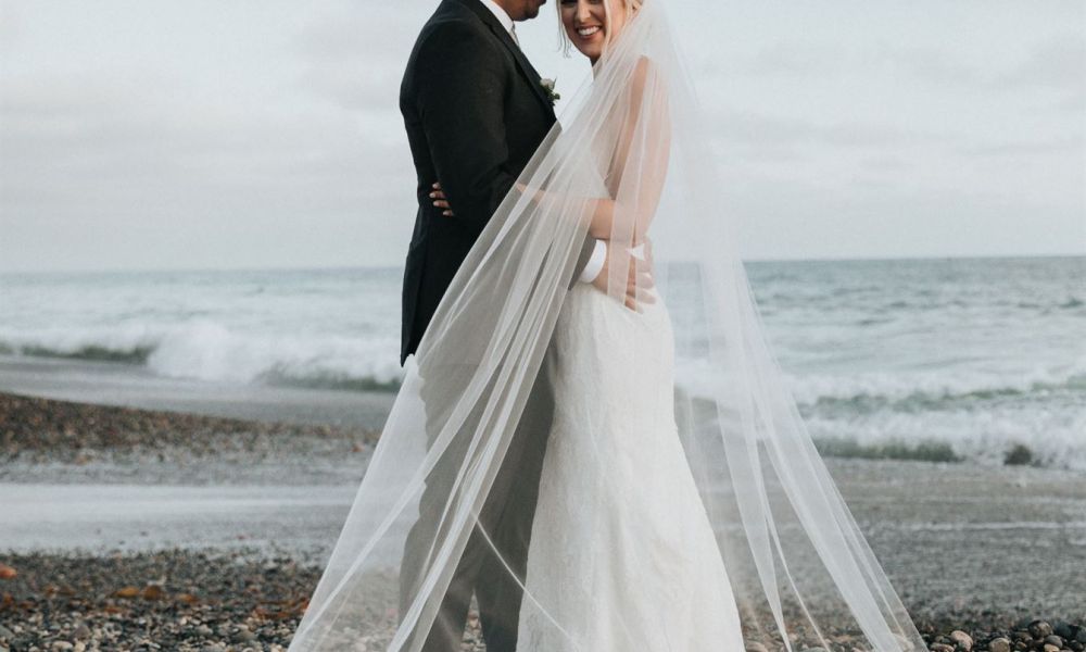 A happy couple stands on a rocky shoreline during their beach wedding. The bride wears a stunning white dress and veil while the groom is dressed in a dark suit. The ocean waves gently lap at the shore, creating a romantic atmosphere as they embrace each other joyfully.