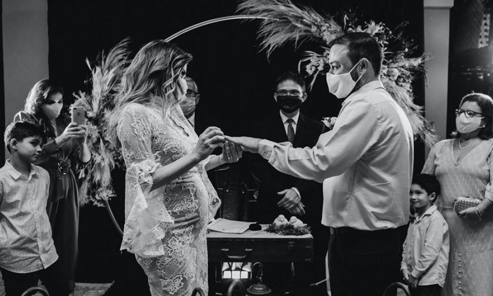 An intimate wedding ceremony scene showing a couple exchanging rings while wearing face masks. The bride looks lovely in her lace dress and the groom is dressed simply in a shirt and trousers. Surrounded by guests who also wear masks, they stand before an officiant under decorative floral arrangements. The ambiance highlights safety protocols during this special occasion while maintaining elegance and warmth with a charming arch behind them.