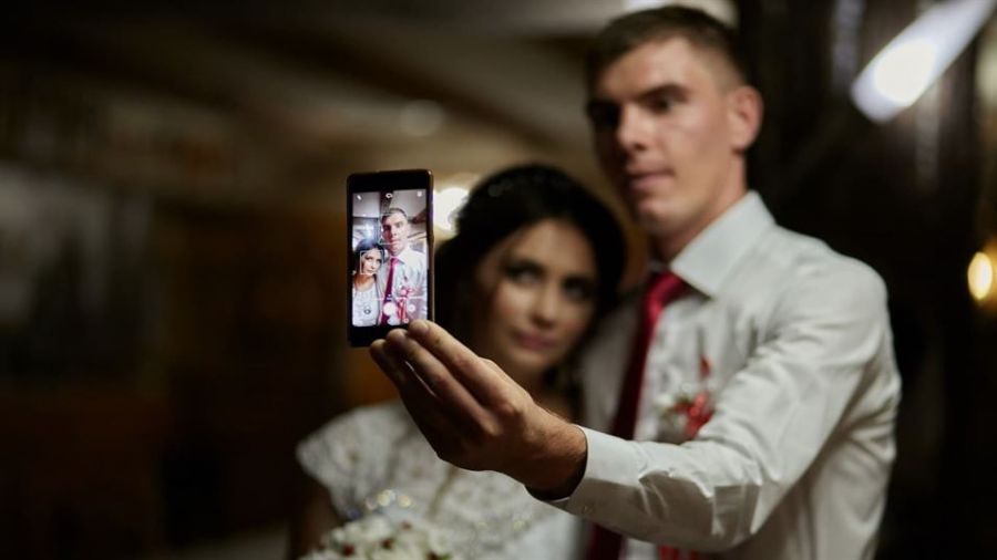 The image features a joyful couple taking a selfie on their wedding day in Dubai. The woman is wearing a beautiful white dress and holding a bouquet while the man dons a white shirt with a red tie. They are close together, smiling at the camera with love in their eyes amidst a warm, softly lit background.