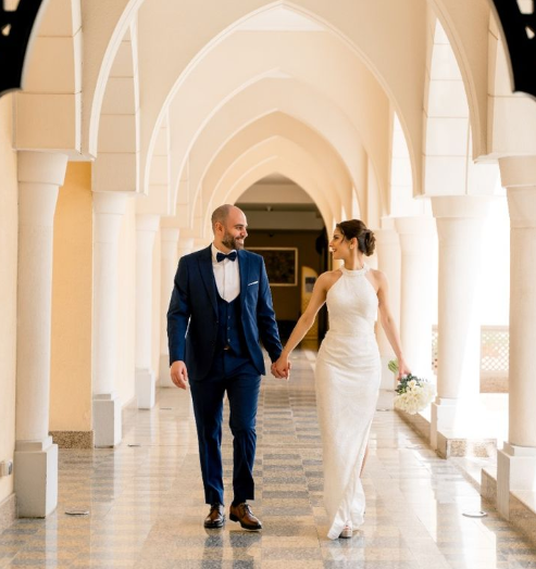 An elegant couple is happily walking hand in hand down a grand corridor. The woman shines in her white gown while the man looks dapper in his blue suit. Intricate architecture surrounds them. Soft light enhances their joyful expressions as they share a loving glance amidst the sophisticated decor. This scene beautifully captures the essence of romance and celebration perfect for any wedding day.