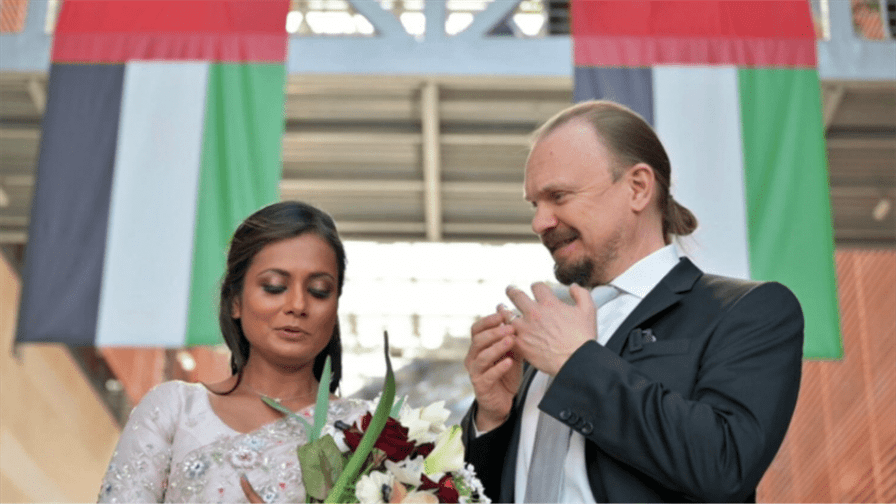 A happy couple stands under two United Arab Emirates flags inside a beautifully decorated venue. The woman holds a bouquet of flowers and wears an elegant dress while the man is in a smart suit. Both share a joyful moment as they look at something together, possibly an engagement ring, surrounded by celebration and love.