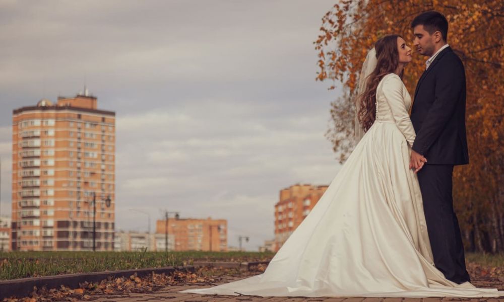 A bride and groom share a loving moment outdoors on a paved path surrounded by vibrant autumn trees. The bride wears a long white gown with a veil while the groom is elegantly dressed in a dark suit. Tall apartment buildings form an urban backdrop against the serene scene filled with golden leaves covering the ground, capturing emotion and warmth on this dreamy day.