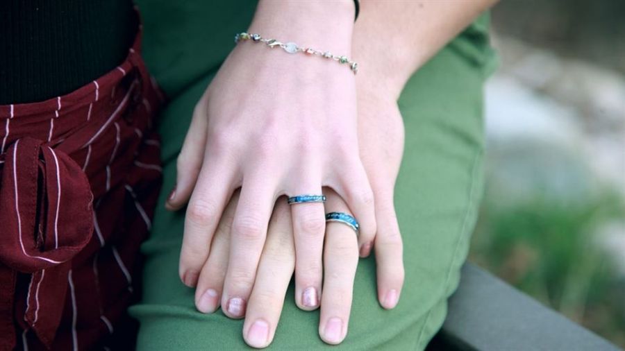 The image features two hands gently resting on top of each other. One hand wears a delicate silver bracelet and a blue stone ring, while the other also has a similar blue stone ring. The background is softly blurred, enhancing the focus on their connection, which symbolizes unity and companionship in love. A maroon, pinstriped fabric is visible near one wrist, indicating casual attire while adding warmth to the scene.
