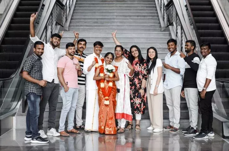 Image of a smiling couple dressed in traditional attire standing at the center of a lively crowd on an escalator as friends cheer them on. The setting is bright and spacious with modern designs surrounding them.