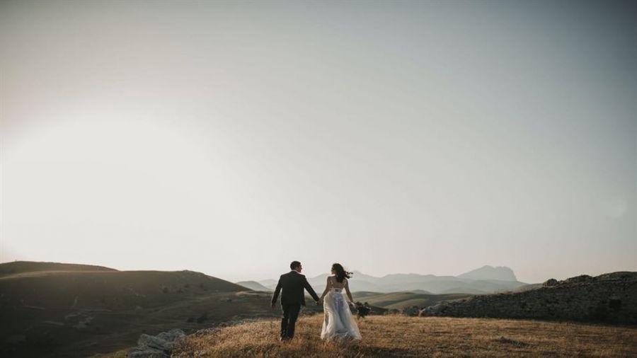 A couple dressed in wedding attire strolls across a serene landscape. The groom wears a dark suit while the bride shines in her flowing white gown. They hold hands, symbolizing love and togetherness against rolling hills and distant mountains under a clear sky. Warm golden light from above adds romance to their moment, creating an enchanting scene that captures the joy of embarking on a new journey together.