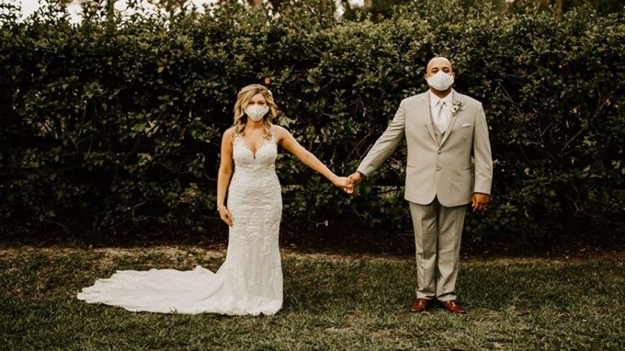 A happy couple stands outside holding hands surrounded by lush greenery. The bride is wearing a beautiful white lace gown while the groom is dressed in a light grey suit. They are both wearing masks, symbolizing love during challenging times. Warm lighting enhances the serene setting, perfect for an intimate wedding ceremony that looks magical and memorable.