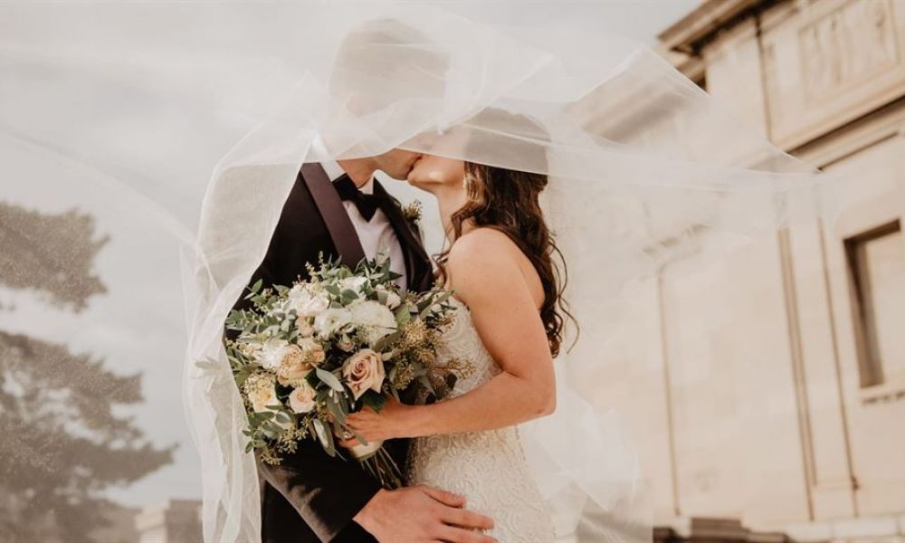 A couple shares a romantic moment under a delicate bridal veil during their outdoor wedding. The groom is wearing a black suit while the bride looks stunning in her white lace gown holding a bouquet of soft-hued flowers. The serene background features trees and elegant architectural elements, creating an inviting atmosphere for their special day.