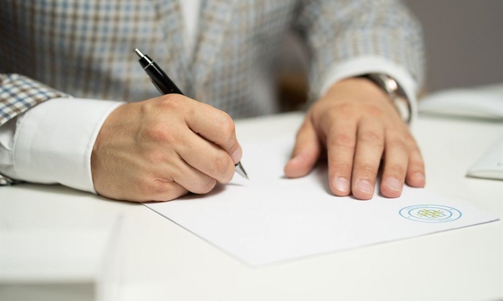 An individual sits at a white table and writes with a black pen on a document that has an official emblem. They wear a stylish checkered blue and white blazer over a light shirt. The scene conveys a formal atmosphere focused on completing marriage-related paperwork, emphasizing the significance of legal documentation in wedding preparations.