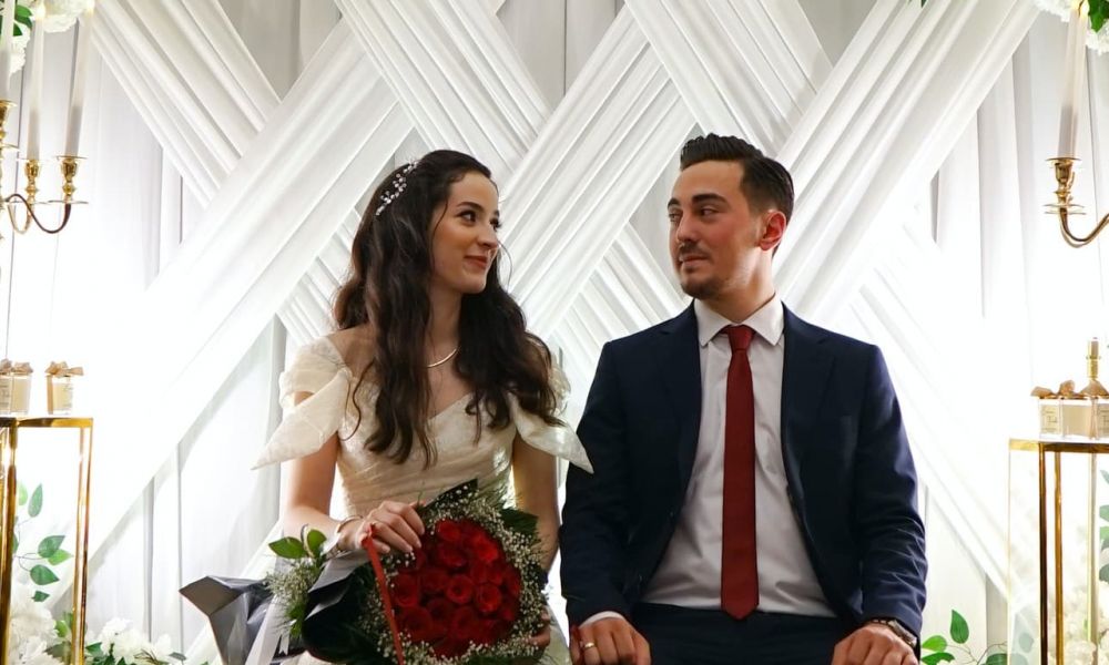 A romantic image of a bride and groom seated together at their wedding ceremony. The bride wears an elegant white dress while holding red roses and has her dark hair styled beautifully. The groom is dressed formally in a navy suit with a bright red tie, as they share happy smiles against a lovely backdrop adorned with greenery and gold candle holders, creating an enchanting atmosphere that reflects love and joy.