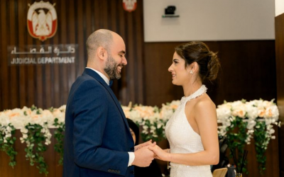 A joyful couple stands hand-in-hand in a formal setting resembling a courtroom. The man wears a dark suit while the woman is in a beautiful white dress. They are smiling with happiness as they celebrate their special day surrounded by elegant decorations.
