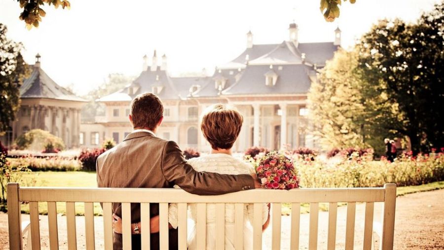 A couple shares a romantic moment on a wooden bench in a picturesque garden. The groom wears a suit while the bride is dressed in white with a vibrant bouquet. They admire a large, charming building surrounded by lush trees and colorful flowers.