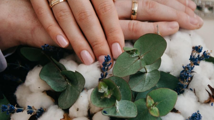 This image shows two hands gently resting on soft white flowers, both wearing beautiful wedding rings. The hands are surrounded by green eucalyptus leaves and delicate blue flowers, all bathed in warm light that creates an atmosphere of love and commitment. The composition symbolizes union and celebration against a natural backdrop, evoking feelings of intimacy and warmth.