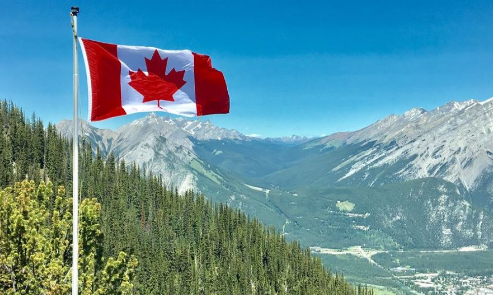 A beautiful image showcasing Canadian wilderness, featuring a waving flag amidst lush green trees and majestic mountains under a clear sky. Lush forests border rugged ranges of snowy peaks in the distance, creating an inviting atmosphere of nature's beauty. Winding roads or rivers snake through verdant valleys below, adding human touch within this stunning landscape filled with tranquility.