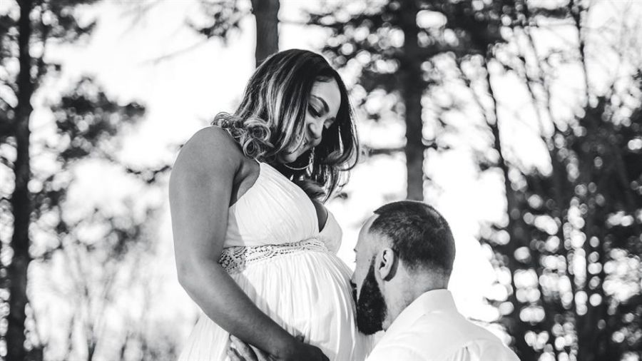 The image shows a loving couple outdoors surrounded by trees. A pregnant woman in a flowing white dress gazes down at her partner who is kneeling or crouching before her. He tenderly kisses her baby bump conveying warmth and affection between them as they anticipate their future together.
