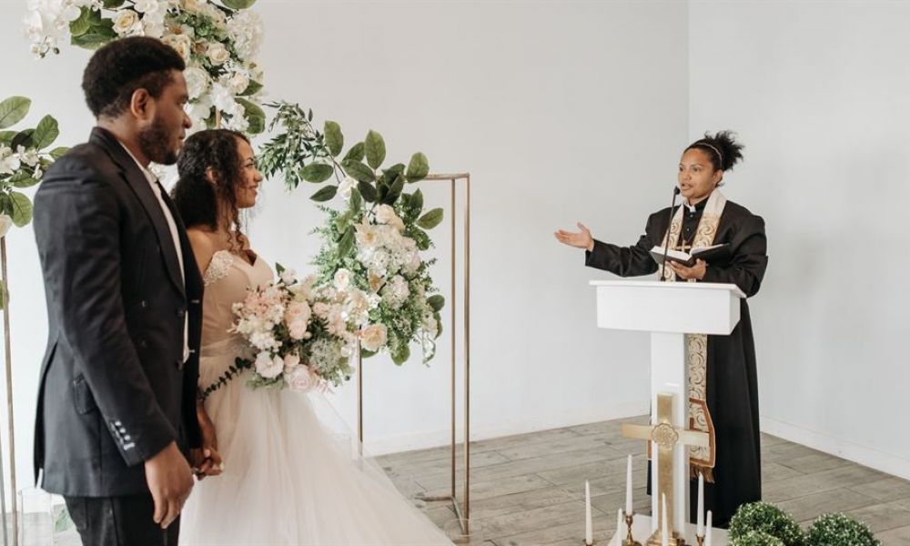 The image shows a joyful couple holding hands during their wedding ceremony in an elegant setting filled with floral arrangements. They appear happy as they stand before an officiant who conducts the ceremony in a warm atmosphere, highlighting love and commitment.