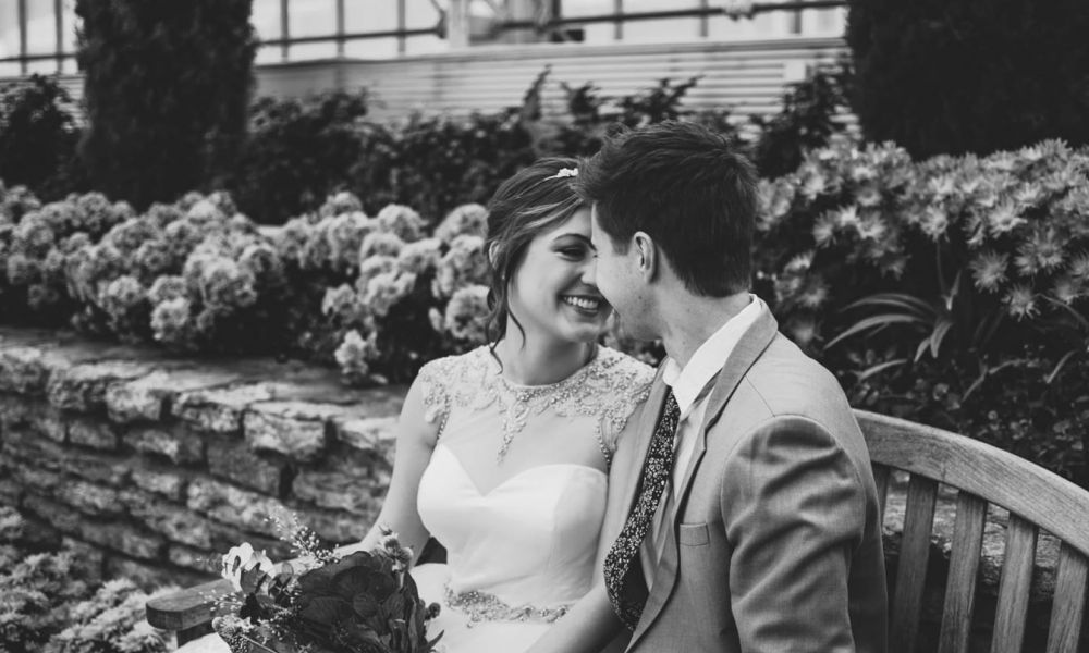 A couple sits closely on a wooden bench surrounded by vibrant green plants and colorful flowers. The bride wears an elegant lace dress and holds a beautiful bouquet, gazing lovingly at her smiling groom, who is dressed in a suit with a patterned tie. They share an intimate moment amid lush greenery, creating a serene atmosphere that encapsulates their happiness as they prepare for their wedding day.