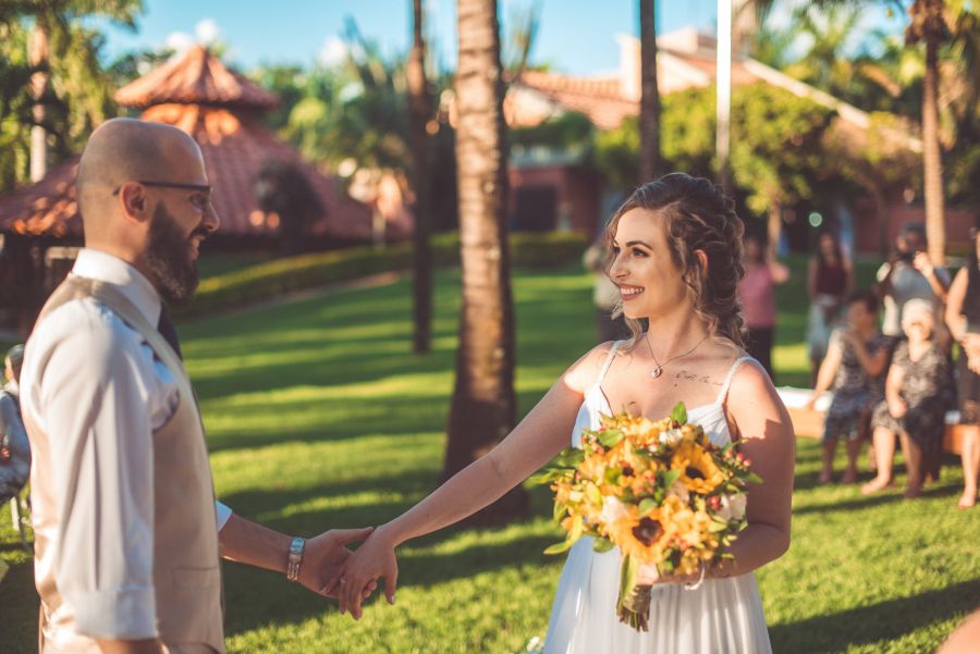 A lovely garden setting captures a couple holding hands during their wedding ceremony in Dubai. The groom wears a white shirt with rolled sleeves and pants while the bride dazzles in a sleeveless white gown with vibrant yellow and orange flowers. Lush greenery surrounds them along with palm trees and guests seated on white chairs celebrating their love on this sunny day under clear blue skies.