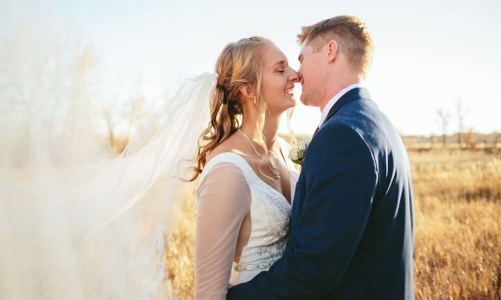 A smiling bride in a flowing white dress embraces her groom in a dark blue suit outdoors. The couple stands in an idyllic sunlit field, conveying love and joy with their warm smiles. Their outfits complement the romantic setting under a clear blue sky, making this moment unforgettable.