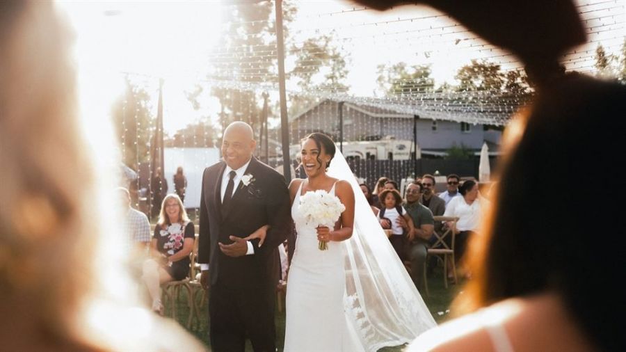 An outdoor wedding scene featuring a bride in a white gown walking down the aisle with her groom beside her. The couple looks happy while surrounded by guests seated on wooden chairs under sunlight filtering through overhead lights. The atmosphere is vibrant and festive, capturing a moment of joy during their wedding ceremony.