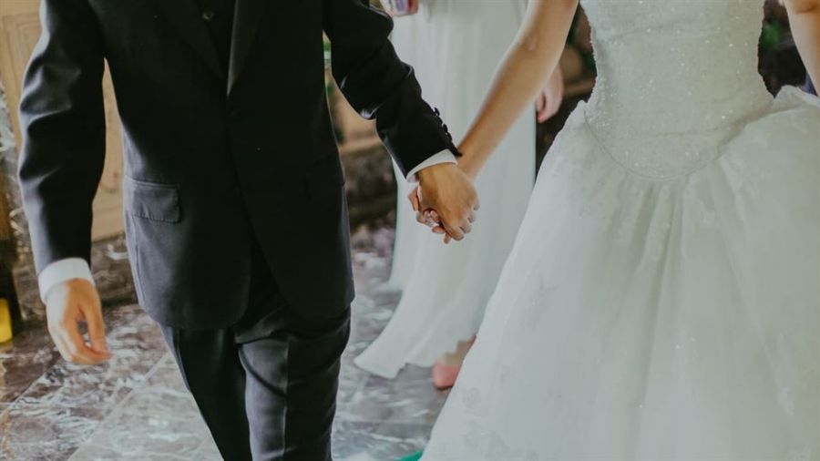 The image shows a happy couple on their wedding day walking hand in hand. The groom is in a classic black suit while the bride wears an elegant white gown with intricate details. They are captured joyfully mid-step against a backdrop of marble flooring and soft lighting that creates an intimate atmosphere celebrating their love on this special occasion.