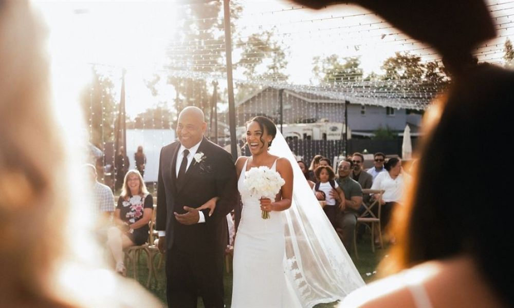 An outdoor wedding scene featuring a bride in a white gown walking down the aisle with her groom beside her. The couple looks happy while surrounded by guests seated on wooden chairs under sunlight filtering through overhead lights. The atmosphere is vibrant and festive, capturing a moment of joy during their wedding ceremony.
